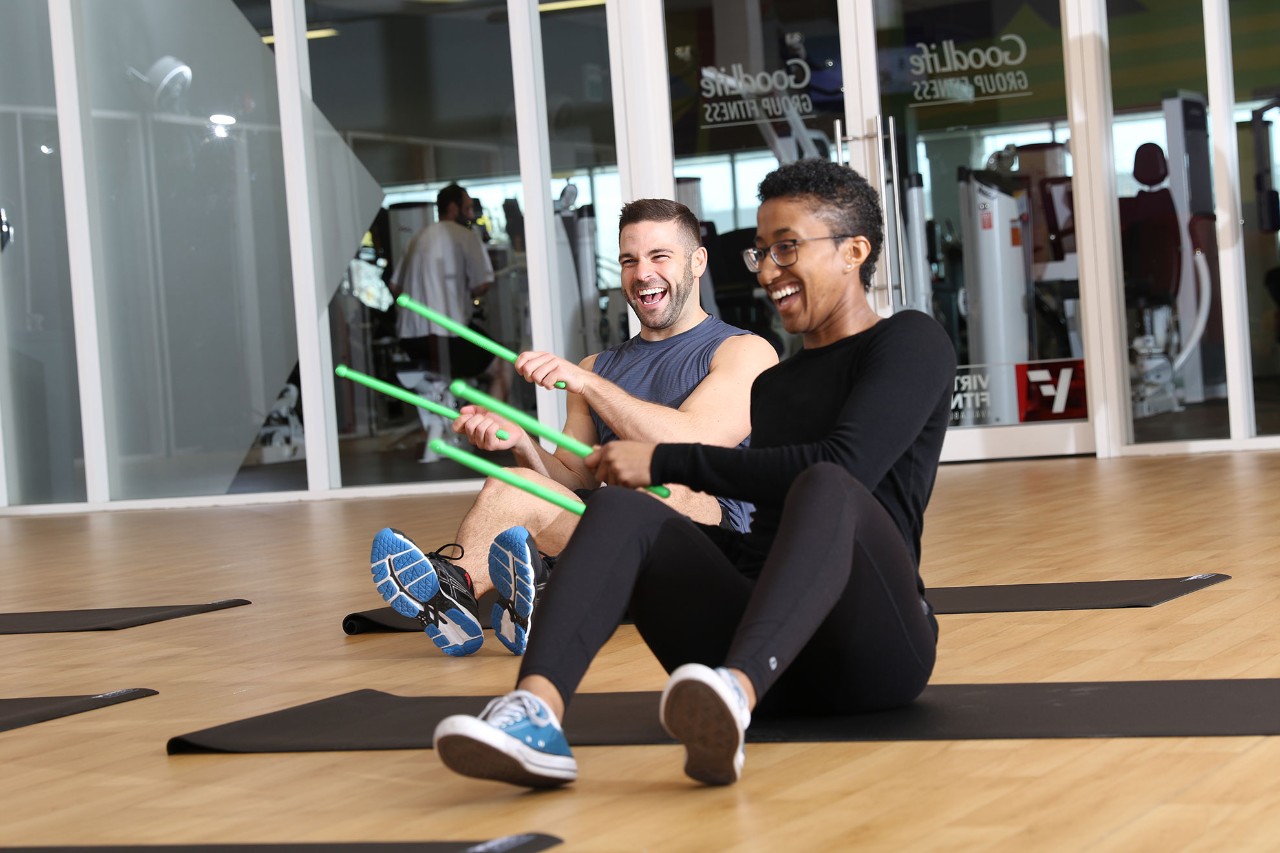 Man and woman with sticks in a GoodLife Pound class, sitting and laughing on yoga mats 
