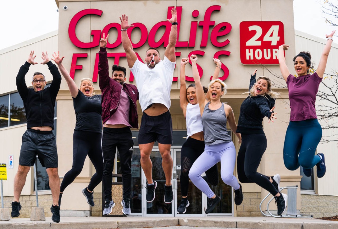 A group of people jumping in celebration in front of the entrance to a GoodLife Fitness club.