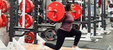 A person in a lunge position and lifting a barbell with large red weights, in a GoodLife Fitness club.