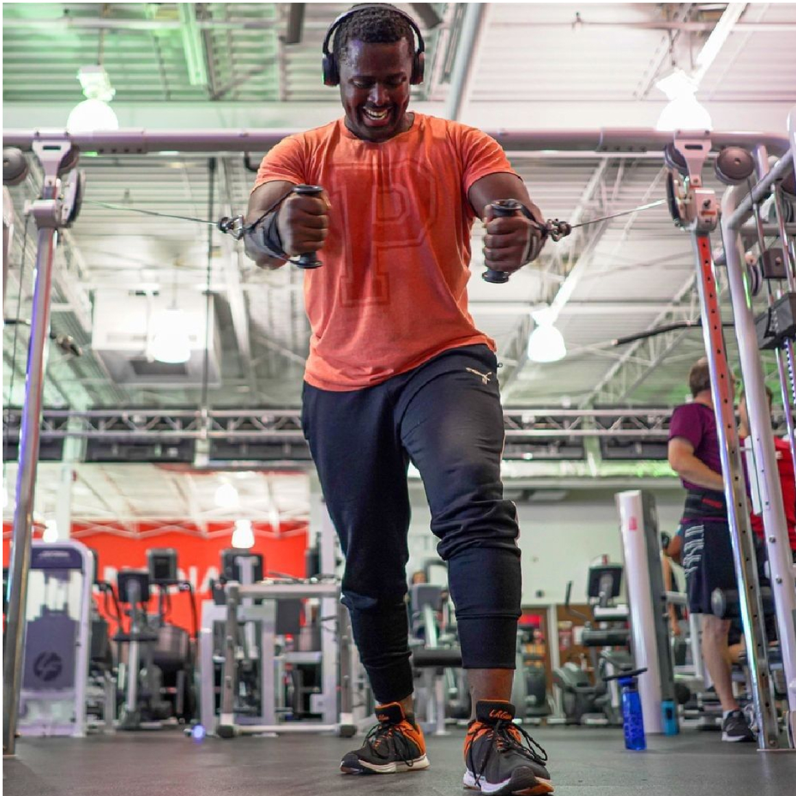 man in orange t-shirt with headphones does a chest press on a pulley machine