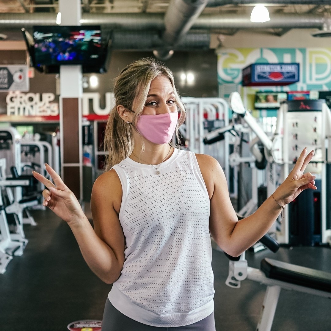 Woman in weight training area wearing a pink face mask looking at camera while making peace signs with her hands. 