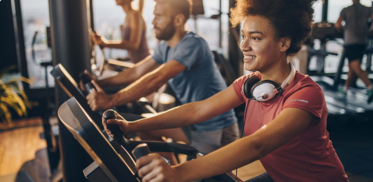 Woman and man work out on stationary bikes with 2 people in the background using cardio machines