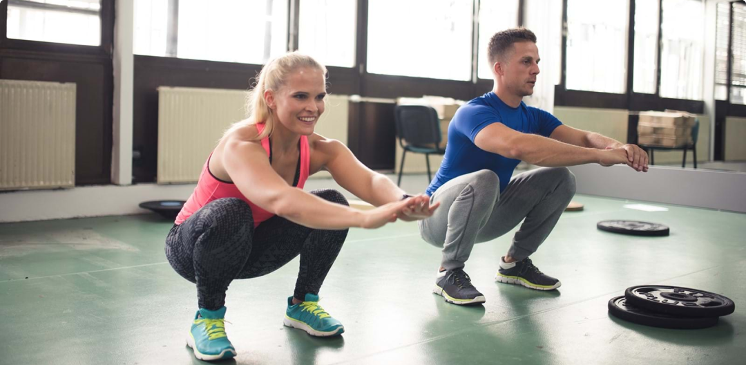 Woman and man both do a deep bodyweight squat in a workout studio with weight plates around them 